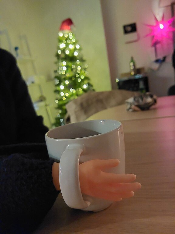 Teil eines Schrottwichtelgeschenks war eine kleine Gummihand, auf diesem Foto hält sie eine Tasse mit Glühwein. Im Hintergrund sieht man einen geschmückten Weihnachtsbaum.