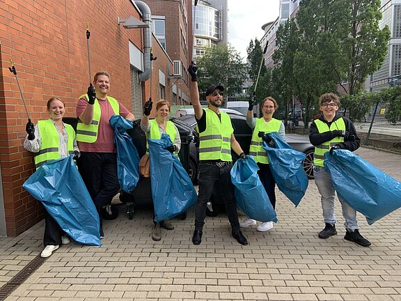 Fröhliches heyday-Team in gelben Warnwesten mit Greifzangen und gefüllten Müllsäcken in den Händen